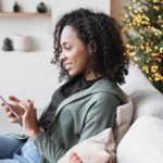 Woman uses her cellphone while sitting by Christmas tree.