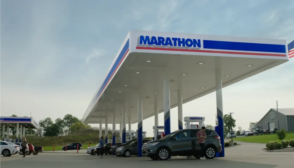 People refuel cars at a Marathon gas station under a large white canopy with the Marathon logo, on a clear day with trees and buildings in the background.