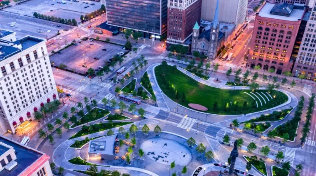 Aerial view of Downtown Cleveland’s city park with green lawns, walking paths, and surrounding streets, bordered by tall buildings and parking lots at dusk.