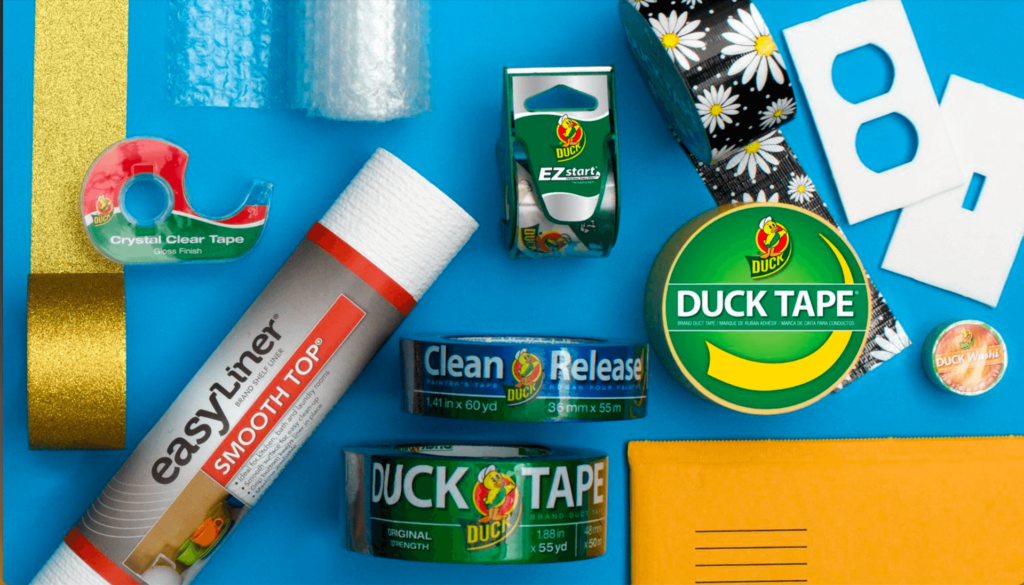 Various Duck brand adhesive products, tapes, a liner roll, scissors, and craft materials arranged on a blue surface.