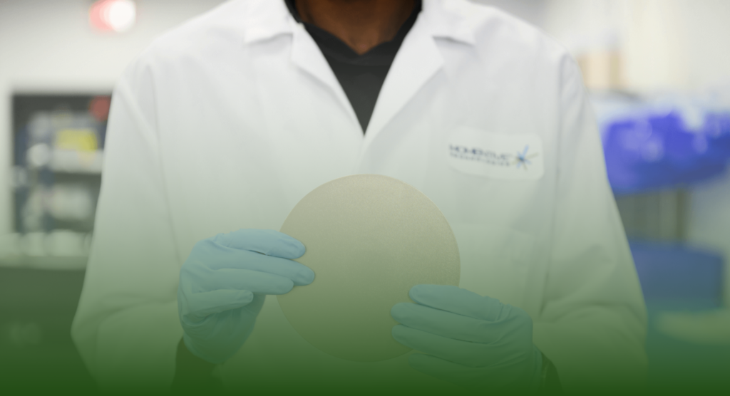 A person wearing a lab coat and blue gloves holds a circular, flat object in a laboratory setting.