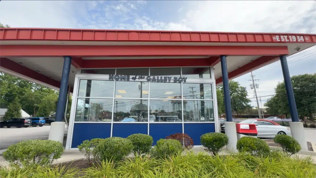 A drive-in restaurant with a red roof, large windows, and sign reading "Home of the Galley Boy," surrounded by neatly trimmed bushes.