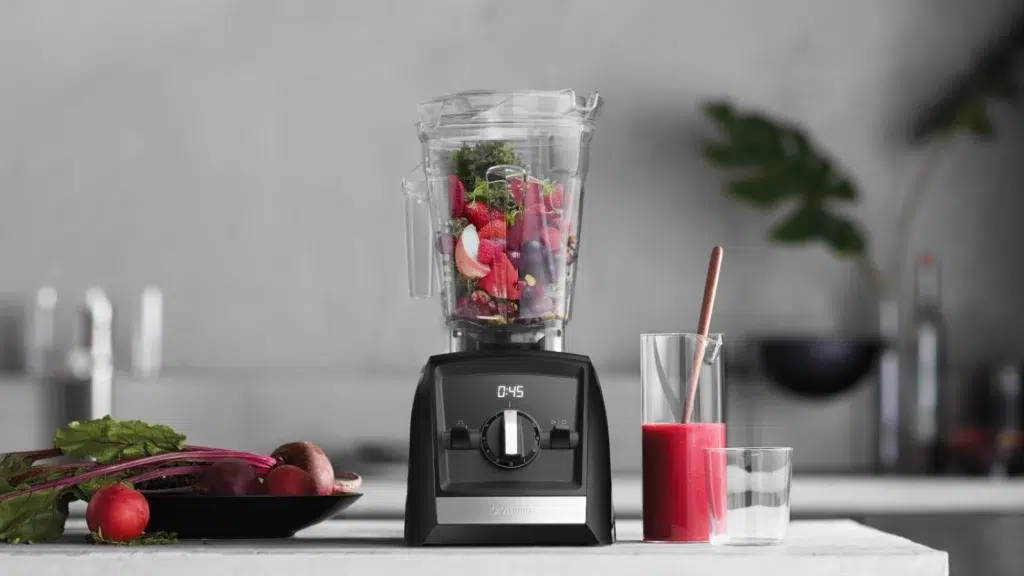 A blender filled with assorted fruits and vegetables stands on a kitchen counter next to a glass of red juice, a plate of produce, and kitchen decor in the background.