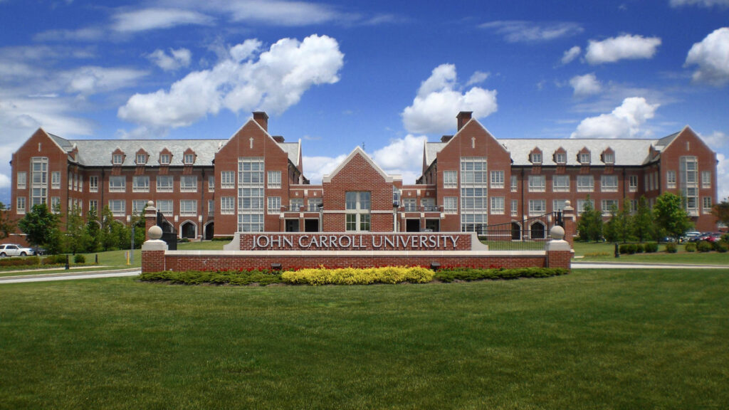 The large brick academic building at John Carroll University stands proudly with its sign in front, surrounded by a lush green lawn and thoughtful landscaping under a bright blue sky dotted with clouds.