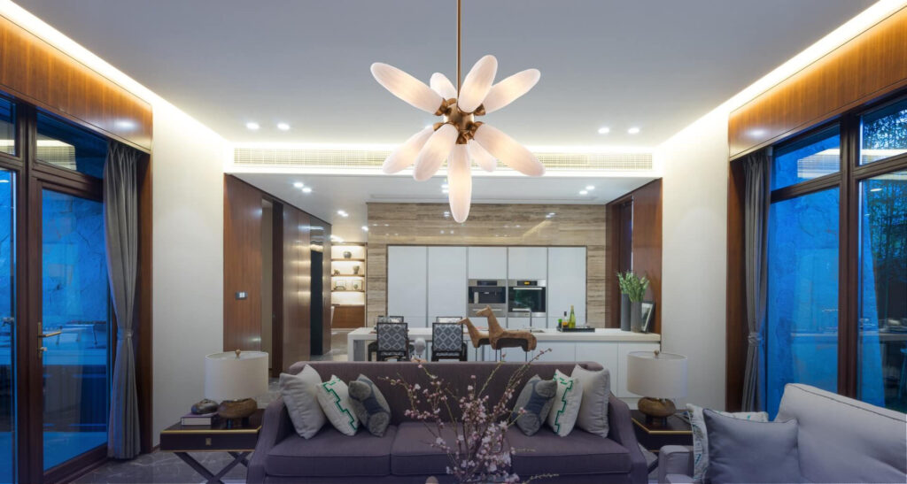 Modern living room and kitchen with wooden accents, a central chandelier, gray sofa, decorative branches, and large windows showing an evening view.