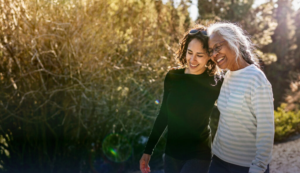 Two women walk arm in arm outdoors on a sunny day, smiling and enjoying each other's company, embodying the supportive spirit of Ohio Guidestone. Trees and sunlight are visible in the background.